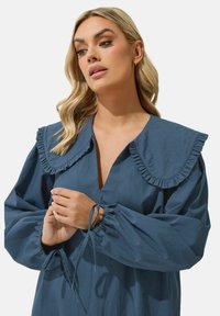 Vestido de algodón azul con un pronunciado escote en V, gran cuello con volantes y mangas abullonadas con puños atados. Textura suave y corte relajado.