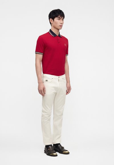 Homme debout portant un polo rouge avec un col noir, un pantalon blanc et des chaussures noires avec des bouts dorés sur fond blanc.