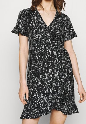 Femme portant une robe portefeuille noire à pois blancs, avec des manches volantées, un ourlet à volants et un collier pendentif en or.