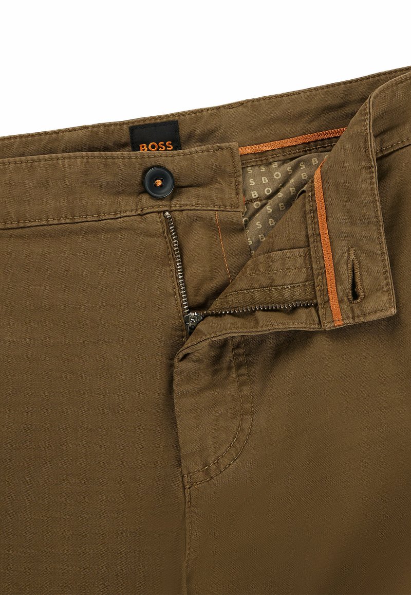 Pantalons marron avec la braguette partiellement zippée, bouton fermé, et doublure intérieure et étiquette de marque visibles au niveau de la taille.