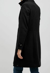 Cappotto in misto lana nero con colletto alto, vestibilità sartoriale e dettagli ai polsini con bottoni. Texture liscia, design di lunghezza media, branding minimal.