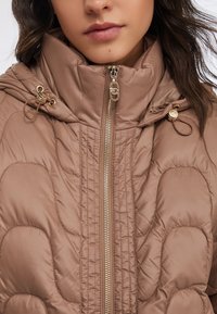 Giacca imbottita marrone chiaro con collo alto, zip dorata e bottoni decorativi. Presenta una texture trapuntata e un design aderente.