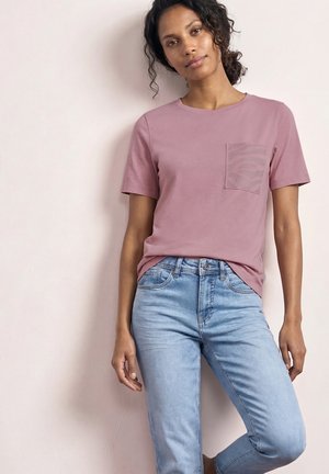 Kvinde iført en mauve kortærmet t-shirt med en tekstureret lomme og lyseblå jeans, stående mod en lysfarvet væg.
