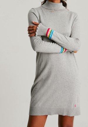 Grå turtleneck sweaterkjole med regnbuefarvede stribede ærmegab og lille pink broderet logo nær kanten, båret med armene krydsede.