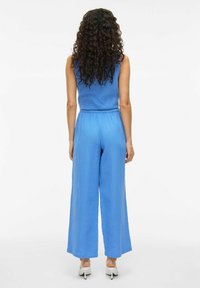 Blauwe jumpsuit met mouwloze top en elastische tailleband, met wijde pijpen en een gestructureerde stof. Draagt met witte hakken.