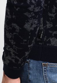 Marineblauer Pullover mit grauem Blumenmuster, gerippten Bündchen und Saum, mit einem Etikett an der Seite; aus weichem Strickmaterial gefertigt.