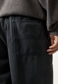 Donkergrijze corduroy broek met een verticale ribbelstructuur, voorzien van een vierkante opgestikte zak aan de rechterzijde. De stof lijkt zacht en duurzaam te zijn.