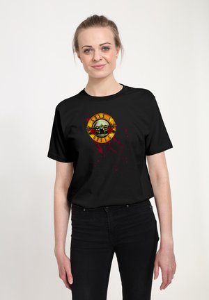 Kvinde med lyst hår sat tilbage iført en sort Guns N' Roses t-shirt og sorte bukser, stående mod en ensfarvet lys baggrund.