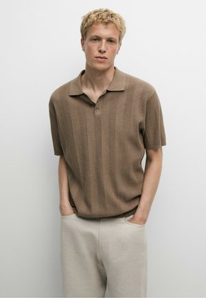 Giovane uomo con capelli biondi e ricci, che indossa una polo marrone a maniche corte in maglia e pantaloni beige chiaro, in piedi con le mani in tasca.