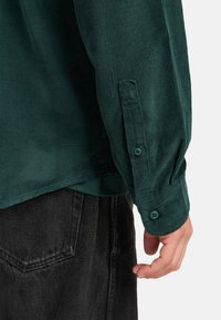 Dark green corduroy shirt with a buttoned cuff, featuring a textured fabric and tailored fit. Black pants partially visible in the background.
