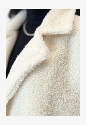 Manteau en polaire couleur crème avec une surface texturée et douce. Présente un col cranté et un gros plan sur le motif en relief du tissu.