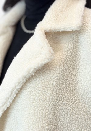 Abrigo de terciopelo sintético color crema con cuello de muesca, que presenta una superficie suave y texturada y un detalle de costura mínima, superpuesto a una blusa negra.