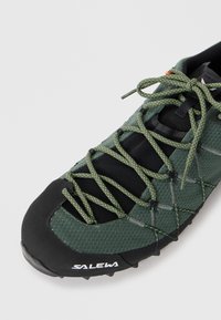 Groene mesh en zwarte rubberen sportschoen met een getextureerde neus en reflecterende groene veters, uitgerust met een veilig vetersysteem en branding aan de zijkant.