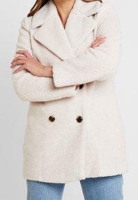 Dwurzędowy płaszcz z miękkiej, teksturowanej kremowej tkaniny, z kołnierzem z klapami i dwoma brązowymi guzikami z przodu.