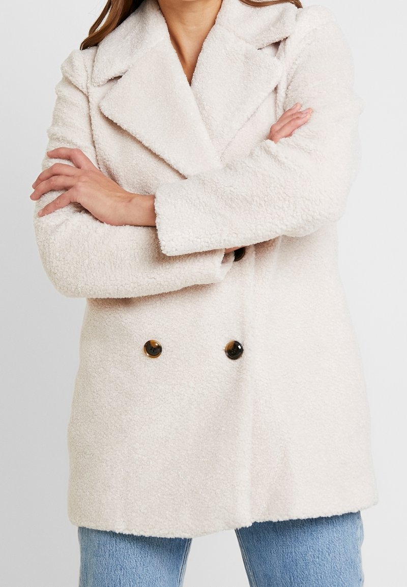 Dwurzędowy płaszcz z miękkiej, teksturowanej kremowej tkaniny, z kołnierzem z klapami i dwoma brązowymi guzikami z przodu.