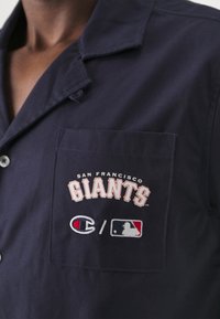 Marineblauw overhemd met knopen en een borstzak. Bevat geborduurd logo van de "San Francisco Giants", Champion-logo en MLB-logo in rood, wit en blauw.
