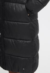 Svart boblejakke med quiltet design, laget av blankt stoff, med en sidelomme og en merkedetalj nær kanten.
