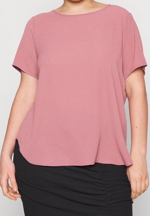 Osoba ubrana w luźną, różowawą bluzkę z krótkim rękawem w kolorze przybrudzonym różu, połączoną z czarną, marszczoną spódnicą, ukazana od szyi do bioder na jednolitym tle.