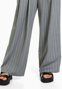 Brede grijze pantalon met streepjes, gedragen met zwarte platform sandalen met een gespsluiting, waarbij de tenen zichtbaar zijn.