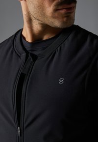 Närbild av en mans nacke och övre bröst som bär en svart zip-up jacka över en svart t-shirt med en liten geometrisk logotyp på jackan.