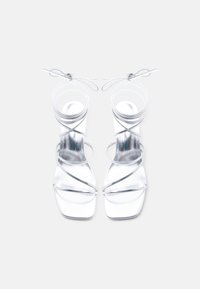 Ζευγάρι ασημί μεταλλικά σανδάλια με λουράκια και ψηλά τακούνια, με τετράγωνες μύτες και δέσιμο στον αστράγαλο, σε λευκό φόντο.