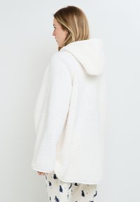 Giacca bianca morbida con cappuccio e superficie testurizzata, caratterizzata da maniche larghe e lunghezza sotto i fianchi, abbinata a pantaloni con pattern.