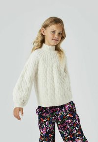 Pull en maille blanche avec un col montant et des manches bouffantes, associé à un pantalon à motif floral coloré. Fond neutre.