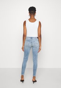 Top blanco sin mangas con bordes en forma de concha, combinado con jeans ajustados azul claro; cuenta con bolsillos traseros y un suave desvanecimiento. Tacones abiertos negros.