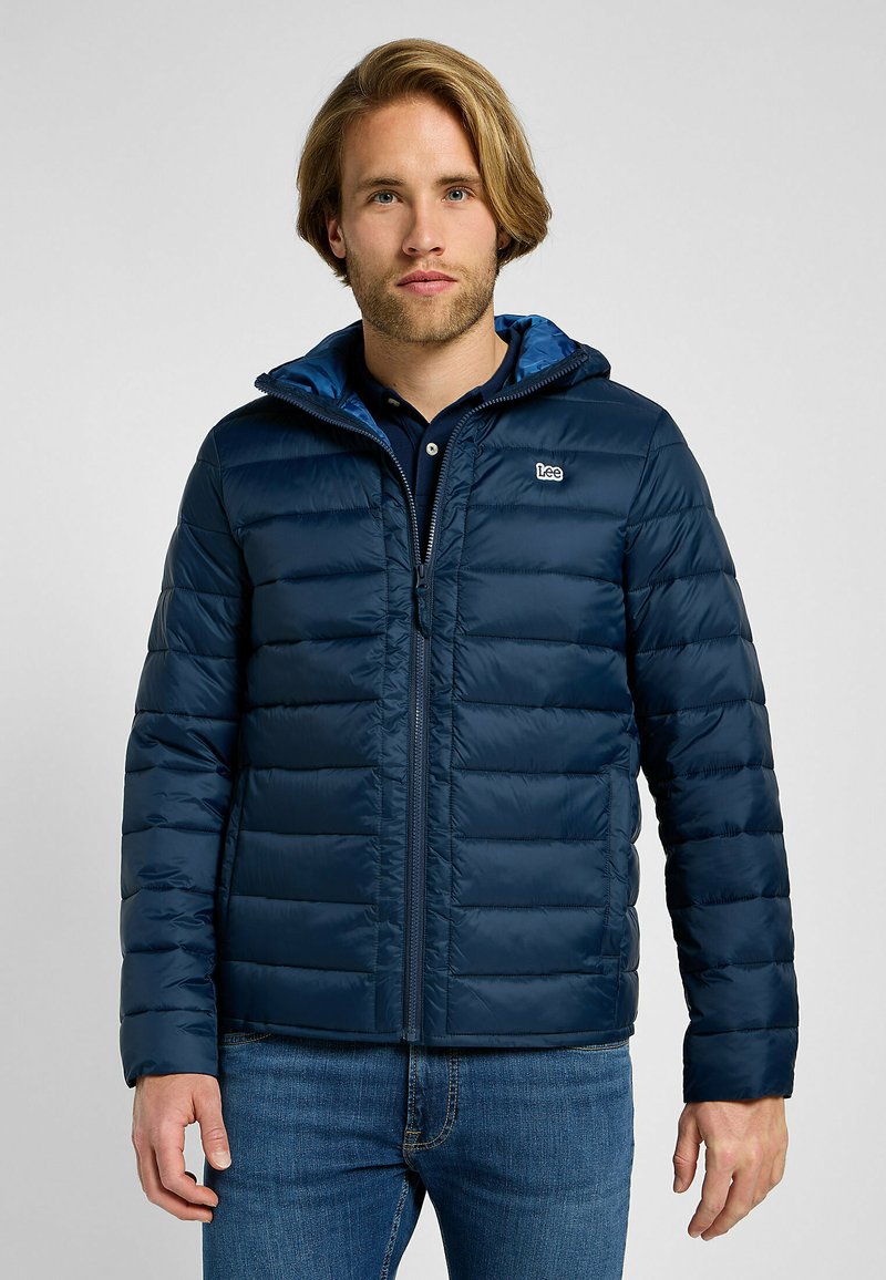 Marineblå puffjakke med glidelås i front, horisontale sømmer og hette, kombinert med en marineblå polo skjorte og denimjeans.