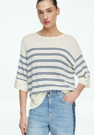 Kobieta w kremowej, luźnej bluzce w poziome paski w kolorze granatowym i niebieskim, w połączeniu z jasnoniebieskimi, wysokim stanem jeansami, z ręką w kieszeni.