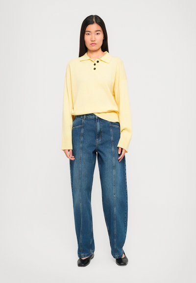 Woman wearing a light yellow long-sleeve polo shirt with black buttons, blue high-waisted jeans, and black low-heeled shoes, standing against white.