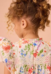 Bloemjurk met pofmouwen, witte stof met kleurrijke bloemenpatronen, kantdetails langs de tailleband, sluiting met knopen aan de achterkant.