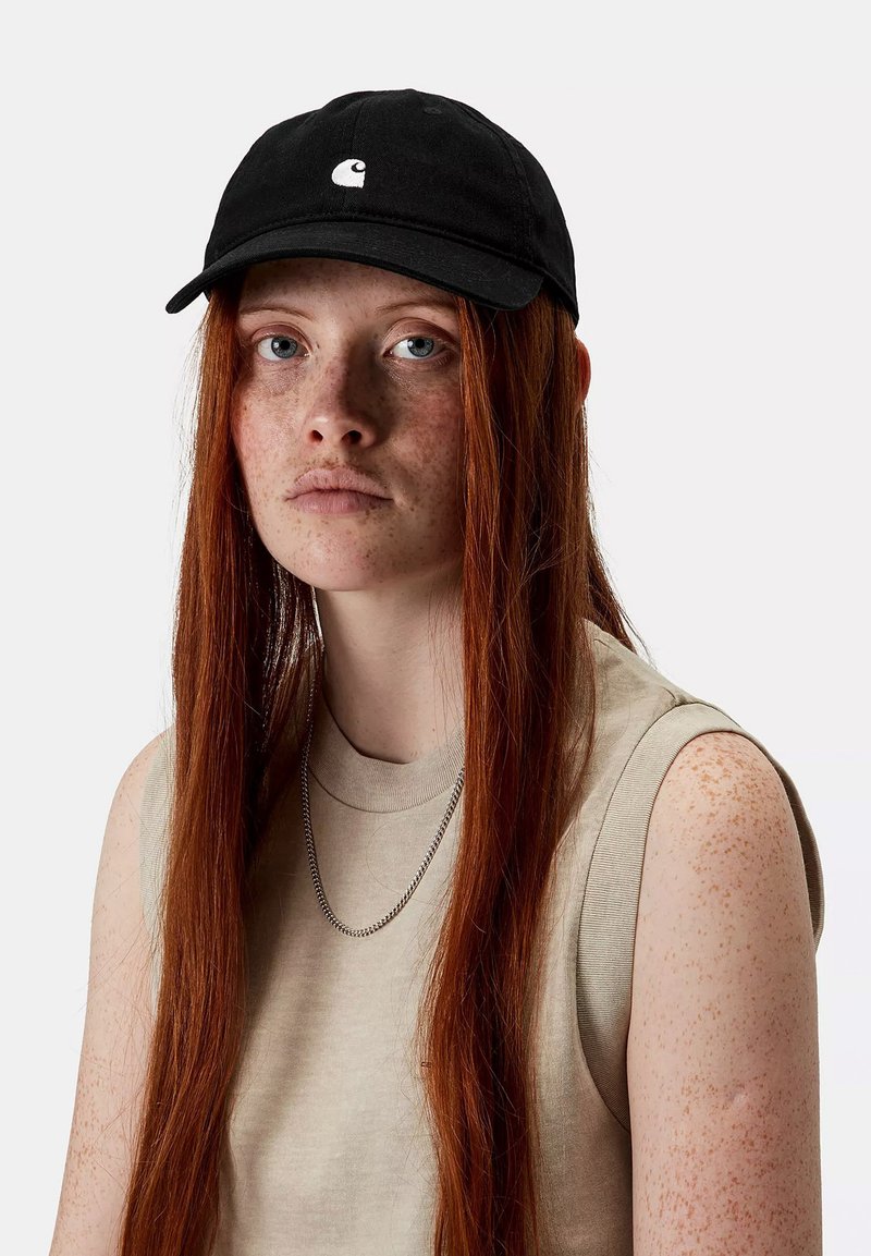 Gorra de béisbol negra con visor curvado y logo blanco, combinada con una blusa sin mangas beige. Cabello rojo largo parcialmente visible. Fondo neutro.