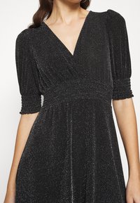 Svart klänning med V-ringning och korta puffärmar, med en samlad midja och glittrig textur över hela.