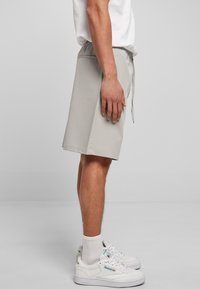 Uomo in piedi di lato che indossa pantaloni corti grigi chiari fino al ginocchio, calze bianche, sneakers bianche Reebok e una maglietta bianca.