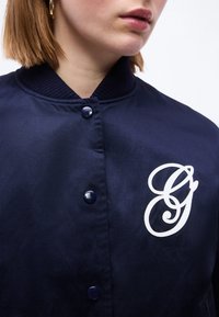 Femme portant une veste bleu marine boutonnée avec une grande lettre cursive blanche "G" sur la poitrine et des boucles d'oreilles créoles dorées.