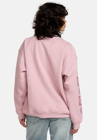 Hanorac roz oversized cu umeri lăsați, textură moale și modele decorative cu soare pe mâneci. Purtați cu blugi de culoare albastru deschis.