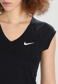 Mujer con una camiseta negra de Nike con escote en V y el logo swoosh blanco en el lado izquierdo del pecho, y cabello rizado visible.