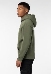Olive green hoodie in soft fabric, featuring a kangaroo pocket and a ribbed hem. Displayed from the side, with a fitted design and adjustable hood.