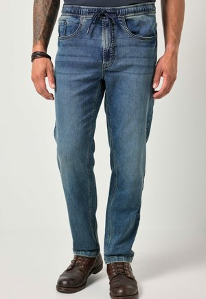 Pantalones de mezclilla en un azul medio con un corte relajado, cintura con cordón, cinco bolsillos y un ligero desgaste en los muslos. Completados con zapatos de cuero marrón.