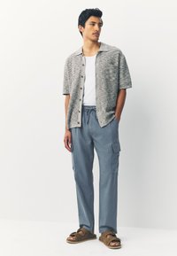 Light gray short-sleeve shirt with buttons, white tank top, blue cargo pants with pockets, and brown sandals with a strap design.