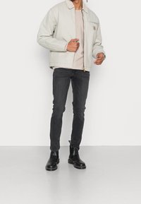 Hellgraue Jacke mit Kragen und Brusttasche, getragen über einem beigen Oberteil und engen schwarzen Jeans, kombiniert mit schwarzen Stiefeletten.