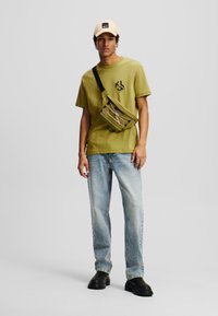 Camiseta verde oliva con logo, pantalones vaqueros de mezclilla azul claro, zapatos negros gruesos y una gorra beige. Bandolera en verde oliva a juego.