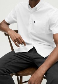 Witte kortekraag button-up shirt met een zak en een merketiket, gecombineerd met zwarte gestructureerde broek. Model zit op een houten stoel.