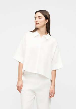 Frau mit glatten braunen Haaren, die ein locker sitzendes weißes kurzärmeliges Hemd mit Kragen und weiße Hose vor einem einfarbigen hellen Hintergrund trägt.