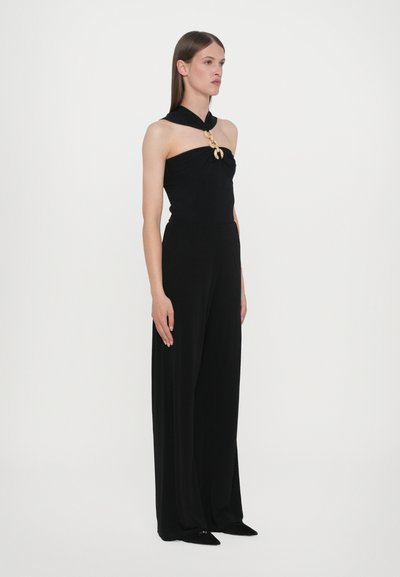 Schwarzer Jumpsuit mit einem enganliegenden, trägerlosen Oberteil mit verdrehtem Design und weiten, fließenden Hosen; geschmückt mit einem goldenen Broschendetail.