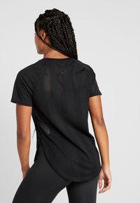 Svart atletisk t-shirt med mesh-textur, korta ärmar och avrundad nederkant. Baksidan visar minimalistisk branding och subtila vertikala ränder.