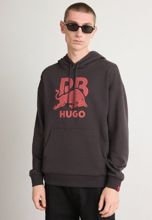 Felpa con cappuccio grigio scuro con un grande logo rosso "PR" e un toro, dotata di tasca frontale e cappuccio regolabile con coulisse. Pantaloni neri e occhiali da sole.