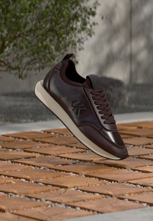 Baskets en cuir marron foncé avec semelle blanche et logo "ck", présentées en l'air au-dessus d'un sol en bois à motifs, avec un arrière-plan flou de plante et de mur.