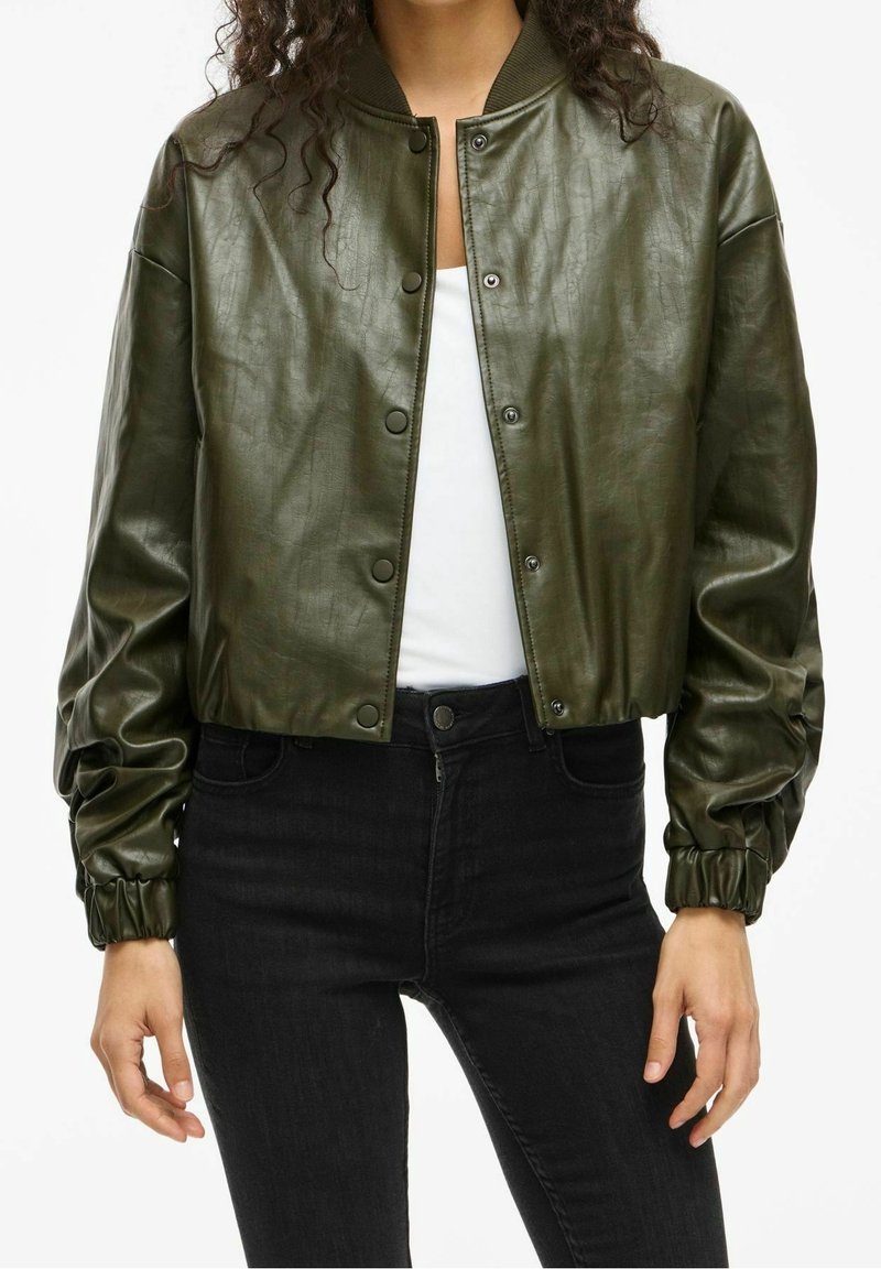 Femme portant une veste bomber courte en cuir synthétique vert olive sur une chemise blanche et un jean noir taille haute, debout, les mains détendues.